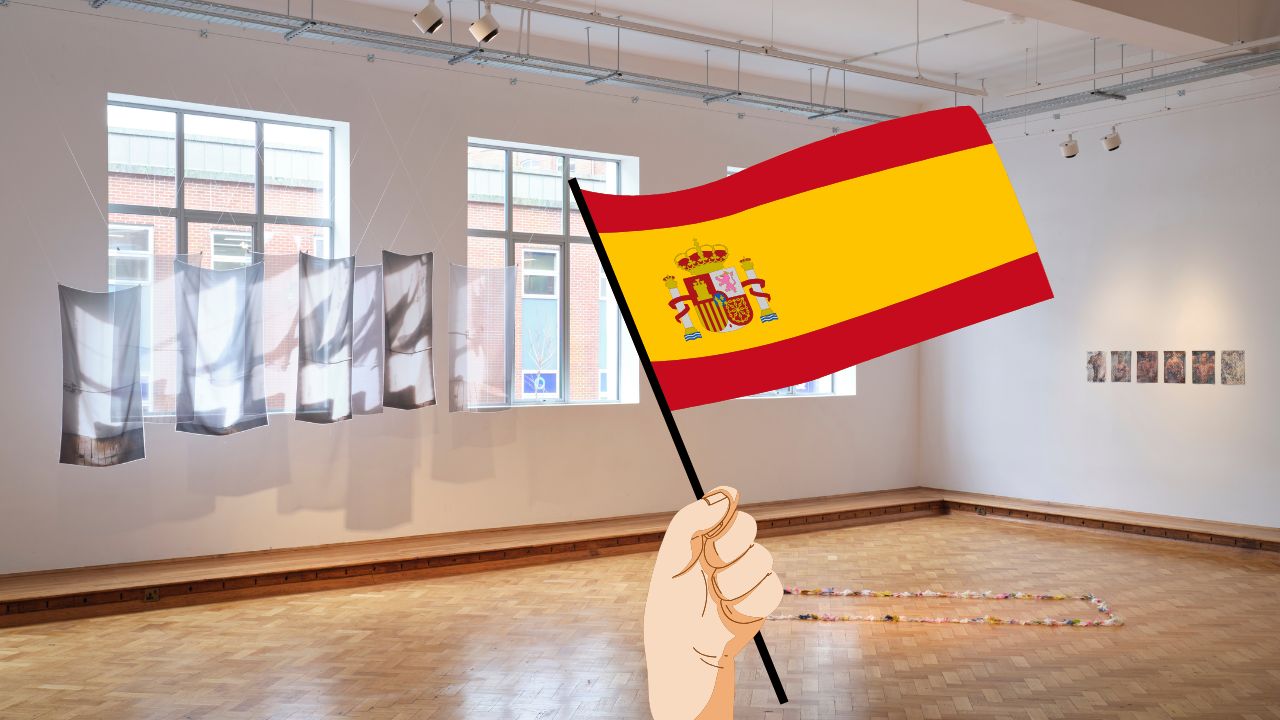 An image of a bright gallery space with wooden floor and white walls. There are three large windows to the left. In front of those windows is a textile artwork of grey and white silk panels hung from the ceiling. To the right is six A4 paper artworks hung in a row on the middle of the wall. An illustration of a hand holding a Spanish flag has been placed in the foreground