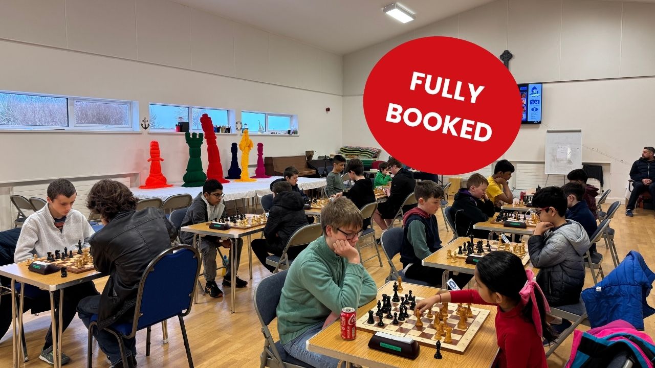 A group of children and teenagers sit at tables in a bright hall, concentrating as they play chess matches. Several boards are set up across the room, with players leaning forward in thought. Colourful oversized chess-piece sculptures line a table along the back wall, and a screen and cross are mounted on the wall.